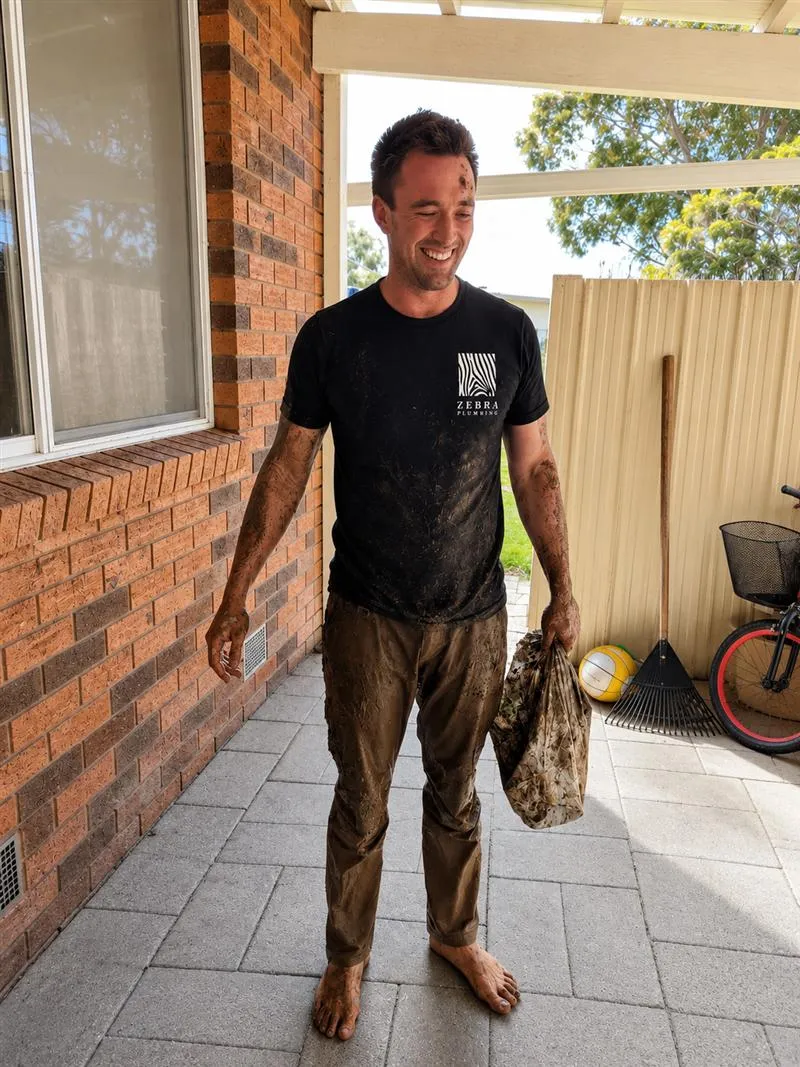 Byron covered in mud after clearing a blocked drain, holding the debris that came out