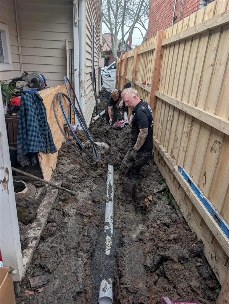 Two Zebra plumbers laying a new PVC sewer line in a muddy trench beside a house