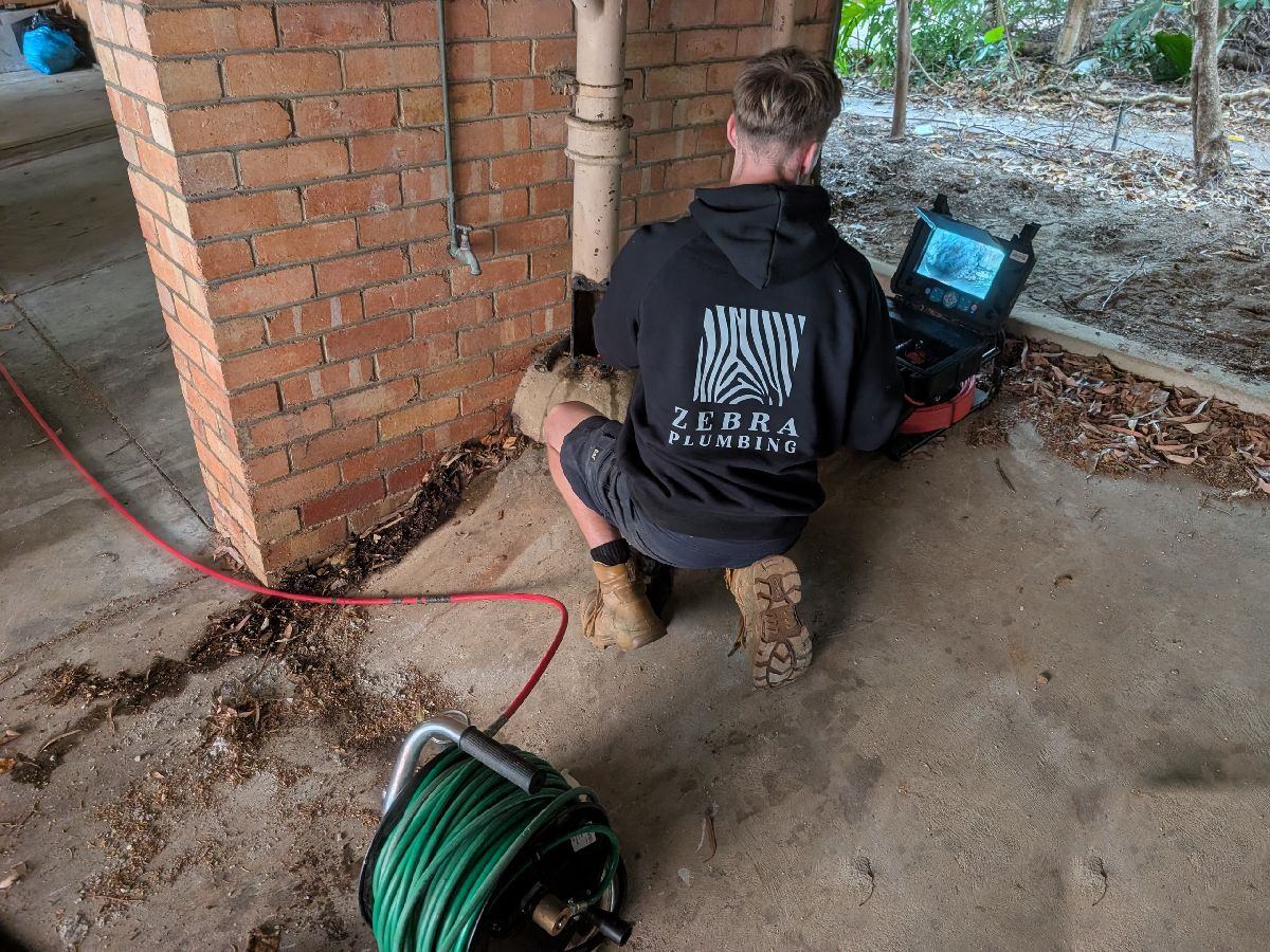 A Zebra plumber crouched at the CCTV monitor beside an outside drain, with the jetter hose reel in the foreground