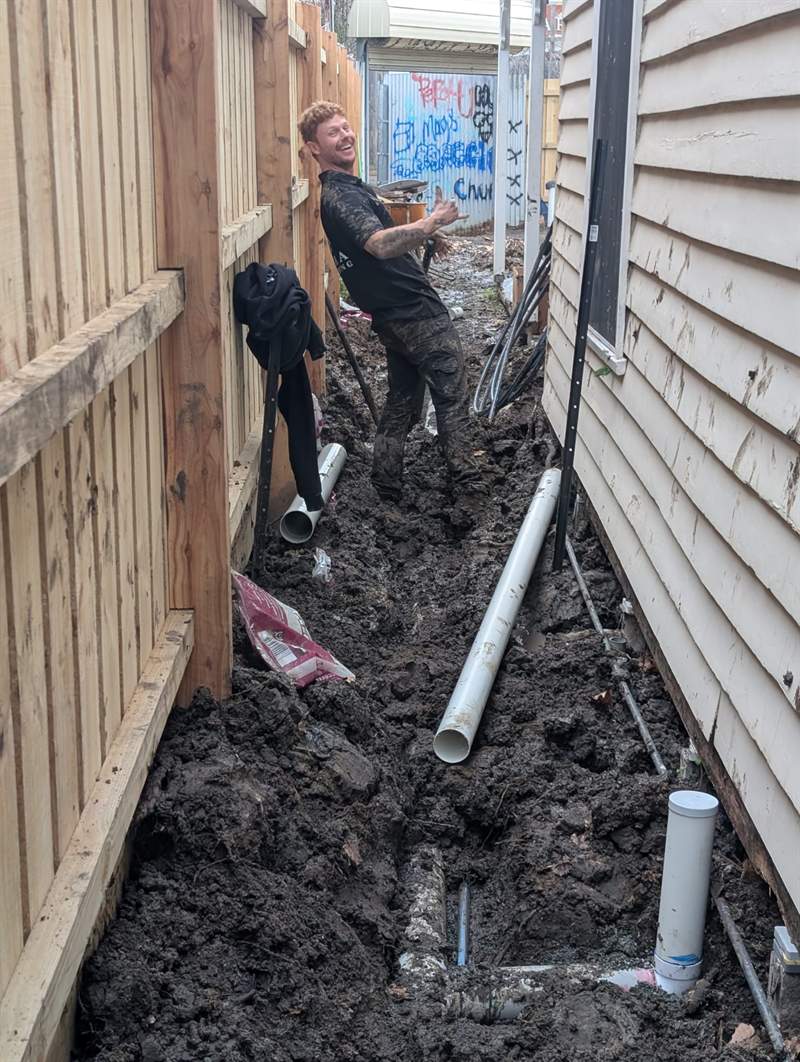 Lucas from Zebra Plumbing covered in mud and grinning beside a freshly dug drainage trench